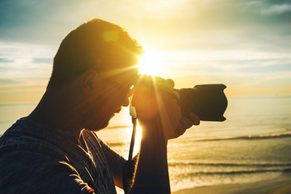 海岸で夕日をバックに一眼レフカメラを構えるプロカメラマン